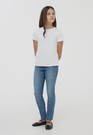 T-shirt bianco a maniche corte con scollatura rotonda, abbinato a jeans skinny azzurri chiari decorati con dettagli in perle bianche. Scarpe nere basse.