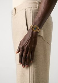 Montre digitale dorée avec un cadran rectangulaire sur une main foncée, associée à un pantalon en laine mélangée texturé beige clair.