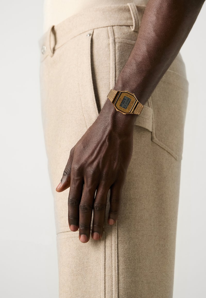Montre digitale dorée avec un cadran rectangulaire sur une main foncée, associée à un pantalon en laine mélangée texturé beige clair.