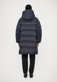 Oseba, oblečena v dolg, temno moder puhovko s kapuco in logotipom EA7 Emporio Armani na hrbtu, črne hlače in črne čevlje, stoji obrnjena stran.