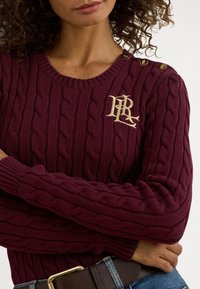 Μπορντό πλεκτό πουλόβερ με σχέδιο κοτσίδας, στρογγυλή λαιμόκοψη, κουμπιά με χρυσό εξάρτημα στον ώμο και χρυσλό λογότυπο στο στήθος.