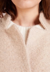 Cappotto beige in lana con una finitura tessuta e textured; presenta un colletto a punte e un morbido interno in maglia color crema. La vista ravvicinata mette in evidenza i dettagli.