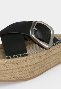 Bride de sandale en cuir noir avec grande boucle argentée sur semelle plateforme beige tressée sur fond blanc.