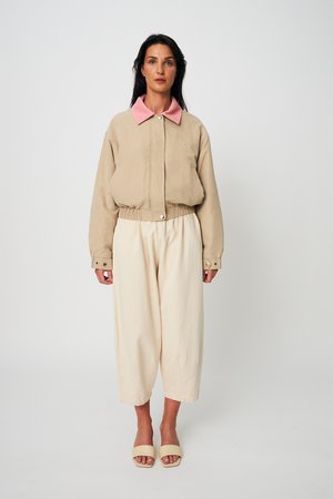 Žena stojící v béžovém zkráceném saku s růžovým límcem, volných krémových kalhotách a béžových sandálech s otevřenou špičkou na bílém pozadí.