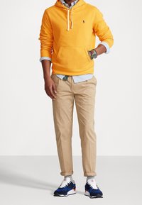 Orange sweatshirt med huva och framficka, beige chinos och blå sneakers. Outfiten inkluderar en ljusblå skjorta och mönstrade handledsarmband.