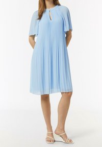 Helles blaues Faltenkleid mit kurzen, flatternden Ärmeln und einem Schlüsselloch-Ausschnitt, aus glattem Stoff hergestellt. Getragen mit weißen, starren Keilsandalen.
