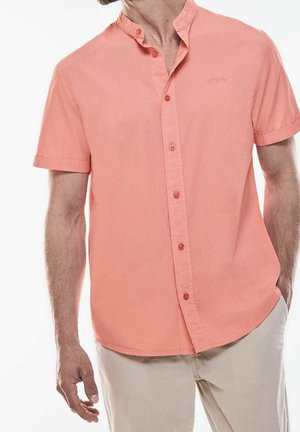 Mężczyzna w koralowej koszuli na guziki z krótkim rękawem i stójką mandaryńską oraz jasnobeżowych spodniach, stojący z rozluźnionymi rękami.