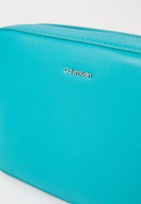 Calvin Klein turquoise cosmetic bag with smooth synthetic material, rectangular shape, and silver logo accent on the front.