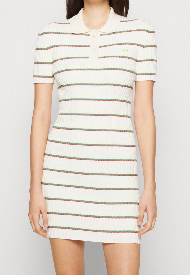 Femme portant une robe polo blanche à manches courtes côtelée avec des rayures horizontales rouges, vertes et jaunes et un petit logo vert sur la poitrine.