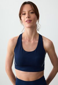 Granatowy biustonosz sportowy z okrągłym dekoltem i wycięciem na plecach. Posiada białe wstawki po bokach oraz gładki, elastyczny materiał zapewniający komfort.