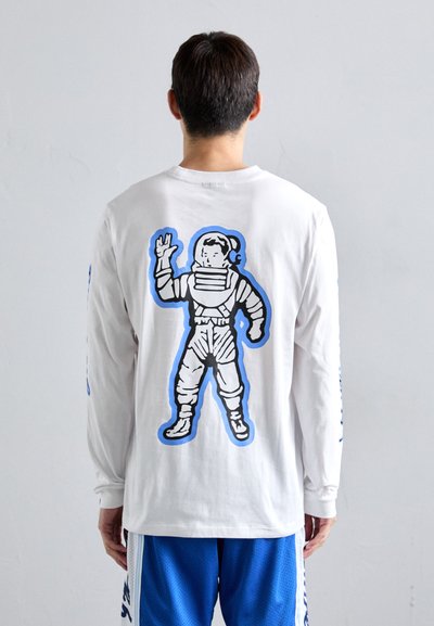 Muškarac nosi bijelu košulju dugih rukava s velikim grafičkim prikazom astronauta u crtežu plavom bojom na leđima, uparenu s plavim sportskim šortsevima.