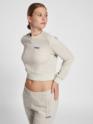 Woman in light gray hummel sweatshirt and sweatpants looks to her left, with one hand on her hip and cropped top showing midriff.