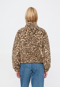Faux fur kurtka w brązowo-beżowy wzór w panterkę, z wysokim kołnierzem i elastycznym pasem, zestawiona z niebieskimi dżinsami.