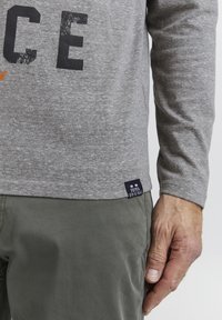 Grå langærmet skjorte med sort og orange tekst, parret med grønne bukser. Et mærke på skjortens afslutning er mærket "F921 Original."