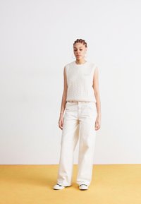 Kremfarbener, ärmelloser Pullover mit Kabelstrickmuster, kombiniert mit lockeren, weißen Hosen und Leinenschuhen, stehend auf einem gelben Boden.