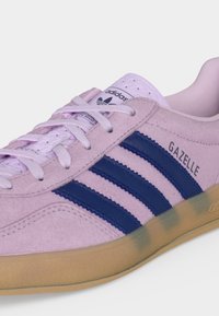 Ανοιχτό μωβ αθλητικό παπούτσι Adidas Gazelle με ναυτικό μωβ ρίγες, λευκά κορδόνια και καουτσούκ σόλα, εμφανίζεται υπό γωνία σε λευκό φόντο.