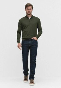 Vihreä vetoketjullinen villapaita, jossa on pitkät hihat, yhdistettynä tummansinisiin farkkuihin ja vaaleanbeigeihin tennareihin. Kangas näyttää pehmeältä ja sileältä.