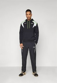 Sweatshirt desportivo preta com detalhes verdes e logótipo, combinada com calças atléticas pretas com detalhes brancos, usada com sapatos pretos e bege.