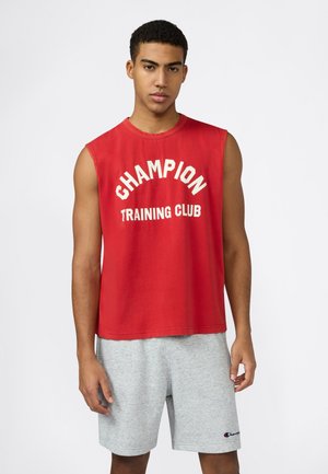 Giovane uomo che indossa una canotta rossa "Champion Training Club" e pantaloncini grigio chiaro Champion, in piedi davanti a uno sfondo bianco.