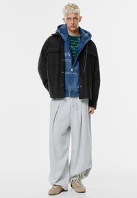 Černá nadměrná bunda vrstvená přes modrou mikinu s kapucí na zip a zelené pruhované tričko, doplněná volnými šedými tepláky a béžovými teniskami.