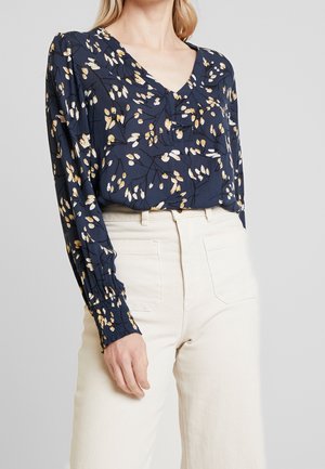 Femme portant un chemisier à manches longues à fleurs bleu marine rentré dans un pantalon taille haute blanc cassé sur un fond uni.