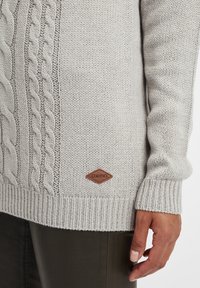 Hellgrauer Strickpullover mit einem Kabelmuster an der Seite, gerippten Bündchen und einem braunen Lederlogo-Patch nahe dem Saum.