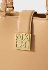Borsa a tracolla in pelle beige con texture, dotata di una placca del logo Armani Exchange dorata attaccata alla tracolla vicino al bordo superiore.