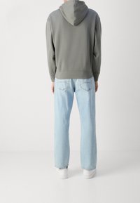 Felpa grigia con cappuccio regolabile, abbinata a jeans in denim azzurro chiaro. Sneakers bianche completano l'outfit visto da dietro.