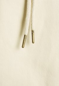 Tissu de sweat à capuche de couleur crème avec une surface texturée. Présente des cordons tressés se terminant par des embouts métalliques. Aucun motif ni logo visible.