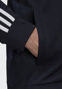 Chaqueta deportiva de color azul marino con acentos de tres rayas blancas en los puños. Cuenta con un bolsillo lateral con un acabado de tela texturizada. Material suave.