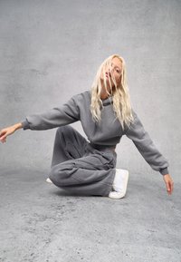 Grijze cropped hoodie en bijpassende sweatpants in zachte stof. Losse pasvorm met elastische tailleband. Model draagt witte sneakers. Detaillering achtergrond.