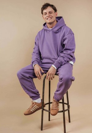 Ein junger Mann sitzt auf einem Metallhocker, trägt einen lila Hoodie, passende Jogginghosen und braune Sneakers und lächelt in die Kamera.