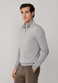 Joven vestido con un suéter de media cremallera gris claro con parches en los codos y pantalones marrones, de pie con una mano en el bolsillo.