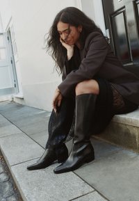 Bottes en cuir noir hauteur genou avec bout pointu et petit talon carré, associées à une jupe en dentelle noire, posées sur une marche en pierre.