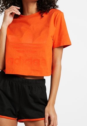 Femme portant un t-shirt court orange Adidas et un short de sport noir avec bordure orange, debout sur un fond blanc.