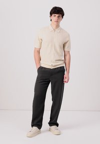 Polo cu mânecă scurtă tricotat în crem, cu un model texturat. Asortat cu pantaloni închiși, lejeri și adidași deschiși. Fundal simplu.