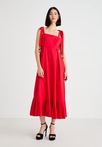 Woman standing in a red midi dress with tied shoulder straps and ruffled hem, wearing black high-heeled sandals, on a plain white background.