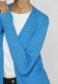 Hellblauer, langärmeliger Cardigan aus weichem Stoff mit gerippten Bündchen und V-Ausschnitt, über einem weißen Oberteil getragen.
