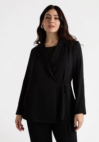 Jachetă neagră, croită, cu design învelit, mâneci lungi și detaliu cu talie legată. Material neted cu un finisaj mat, combinată cu un top negru dedesubt.