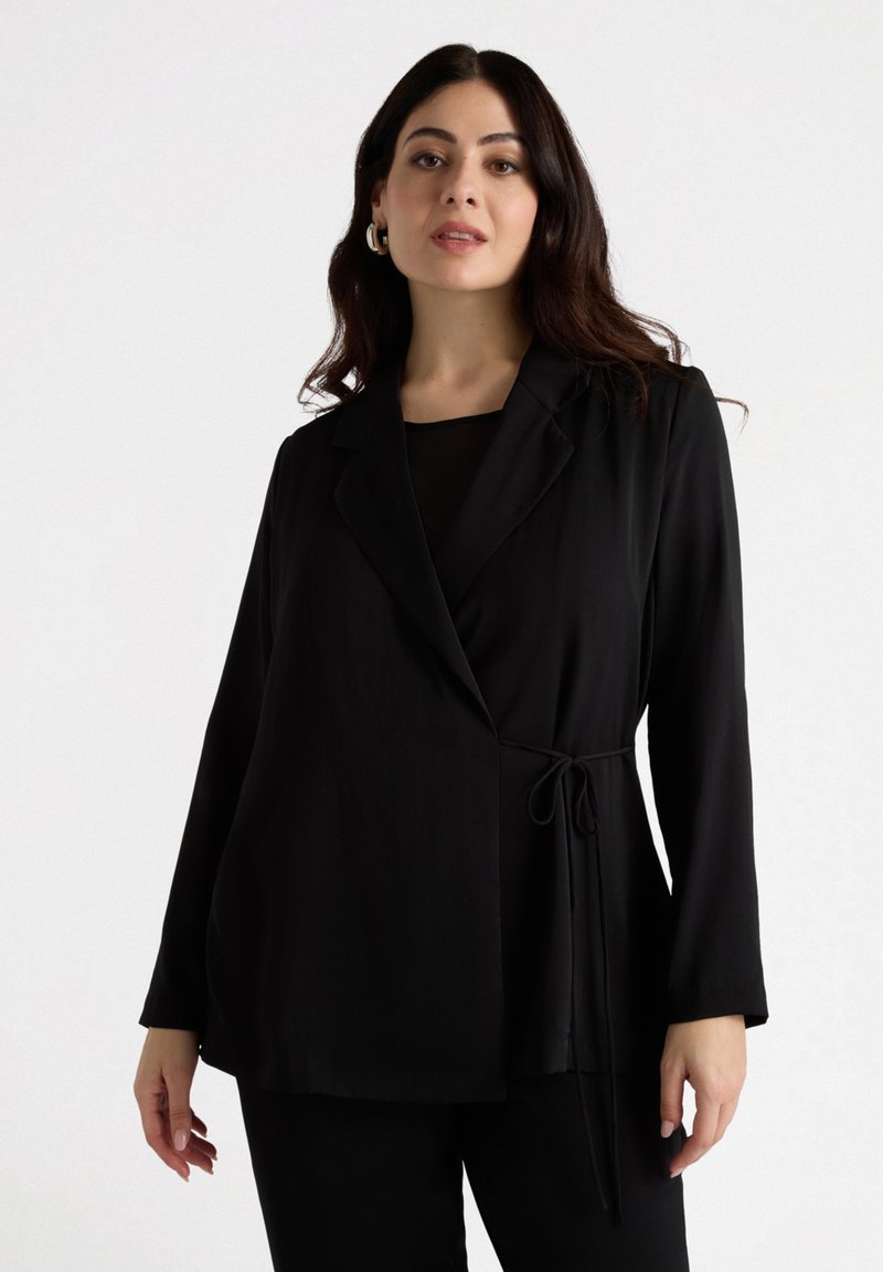 Jachetă neagră, croită, cu design învelit, mâneci lungi și detaliu cu talie legată. Material neted cu un finisaj mat, combinată cu un top negru dedesubt.