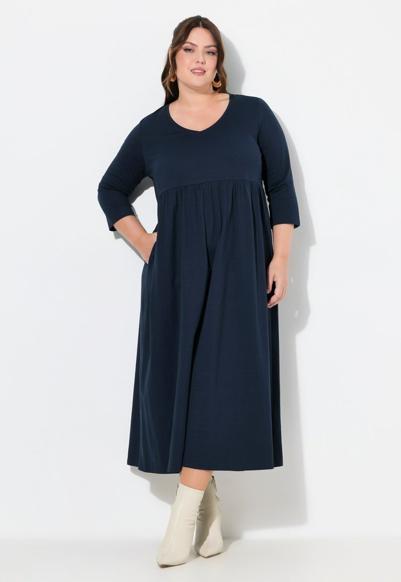 Marineblauwe jurk tot de knie met een V-hals, 3/4 mouwen en een geplooide taille. Gemaakt van zachte stof; heeft zijzakken. Gecombineerd met witte enkellaarsjes.