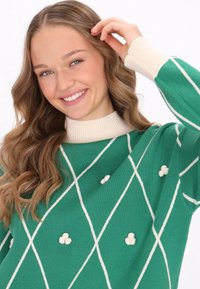 Maglione verde lavorato a maglia con un colletto rigato color crema, caratterizzato da un motivo a rombi e dettagli testurizzati con pompon bianchi sparsi.