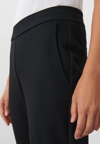 Svarta texturerade byxor med flat midja, sidofickor och en strömlinjeformad silhuett; tyget visar subtil diagonal ribbingdetalj.