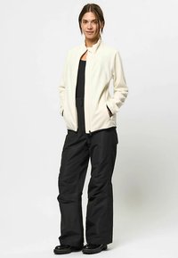White fleece jacket with a high collar, front pockets, paired with black waterproof pants and black boots, on a neutral background.