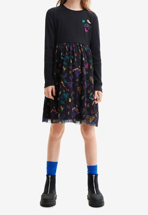 Schwarzes, geripptes Langarmkleid mit einem bunten Herzen und einem Wortmuster am Rock. Kombiniert mit schwarzen Stiefeln mit Reißverschluss und blauen Knöchelsocken.