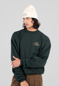 Uomo con capelli ricci scuri che indossa un cappello di maglia crema, una felpa verde scuro con logo e pantaloni marroni, che guarda di lato con le braccia incrociate.