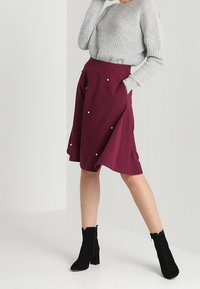 Burgunderfarbene knielange Röcke mit seitlichen Taschen, geschmückt mit dekorativen weißen Perlen. Kombiniert mit einem grauen Strickpullover und schwarzen Schnürstiefeletten.