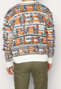 Mehrfarbiger Pullover mit einer Mischung aus Orange, Schwarz und Weiß, ausgestattet mit fransigen Texturen und geometrischen Mustern, sowie gerippten Bündchen und Saum.