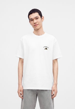 Young man wearing a white T-shirt with a small graphic and text on the left chest, paired with light gray pants, against a plain background.
