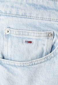 Světlá modrá denimová látka s viditelnou prošitou kapsou. Obsahuje malé vyšívané logo v červené, bílé a modré barvě a stříbrný druk.
